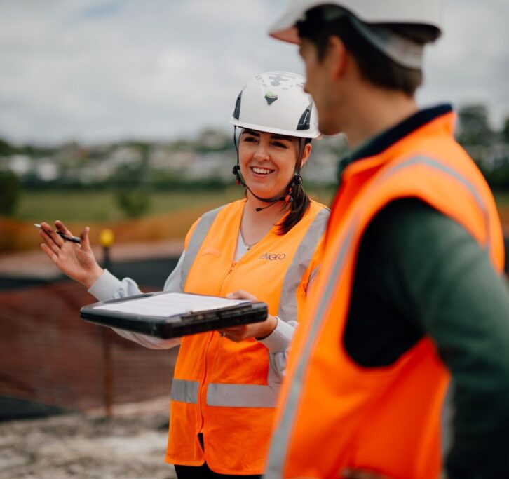 Woman and man in hardhats and safety jackets conversing on a project outdoors.