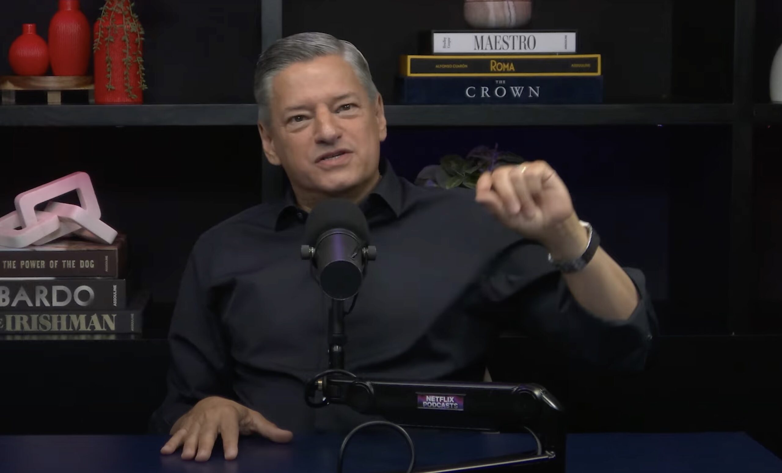 A man in a dark shirt speaks into a microphone while gesturing with his hand. He sits at a table with Netflix-themed books stacked behind him and dark shelves displaying books and decor.
