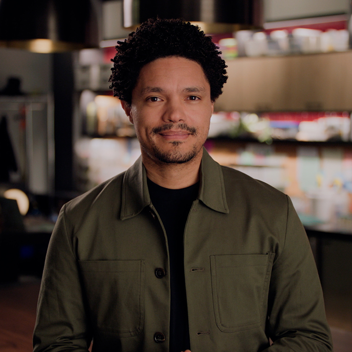 A person with short curly hair, wearing a green jacket, stands in a room with shelves and various objects in the background.