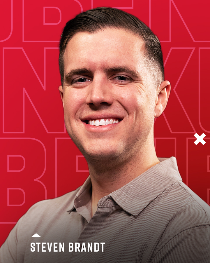 A man with short brown hair smiles at the camera, wearing a light gray collared shirt. The red background features large faint letters and the name "Steven Brandt" at the bottom.
