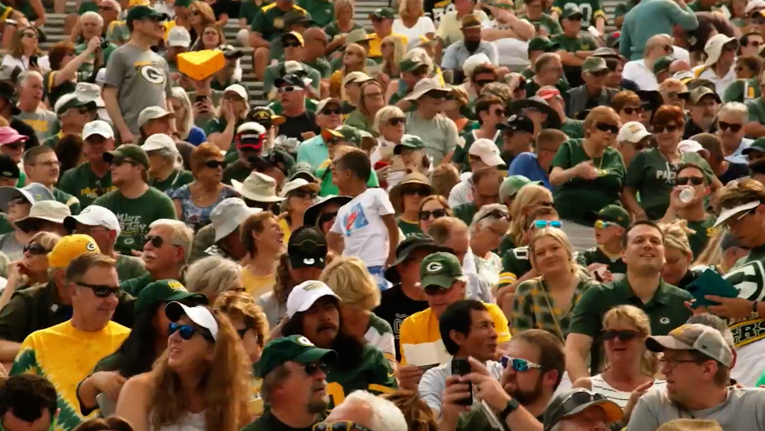 A large crowd of people, many wearing green and yellow sports apparel and hats, sit closely together in stadium seating, watching an event.