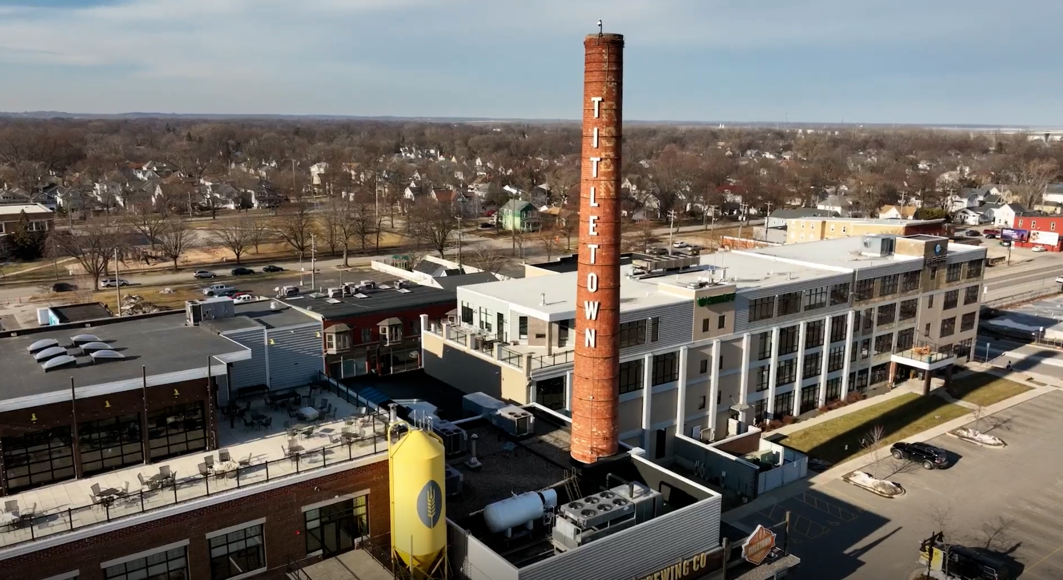 A tall brick smokestack labeled "TITLETOWN" rises above an industrial building complex in a suburban area with houses and trees in the background.