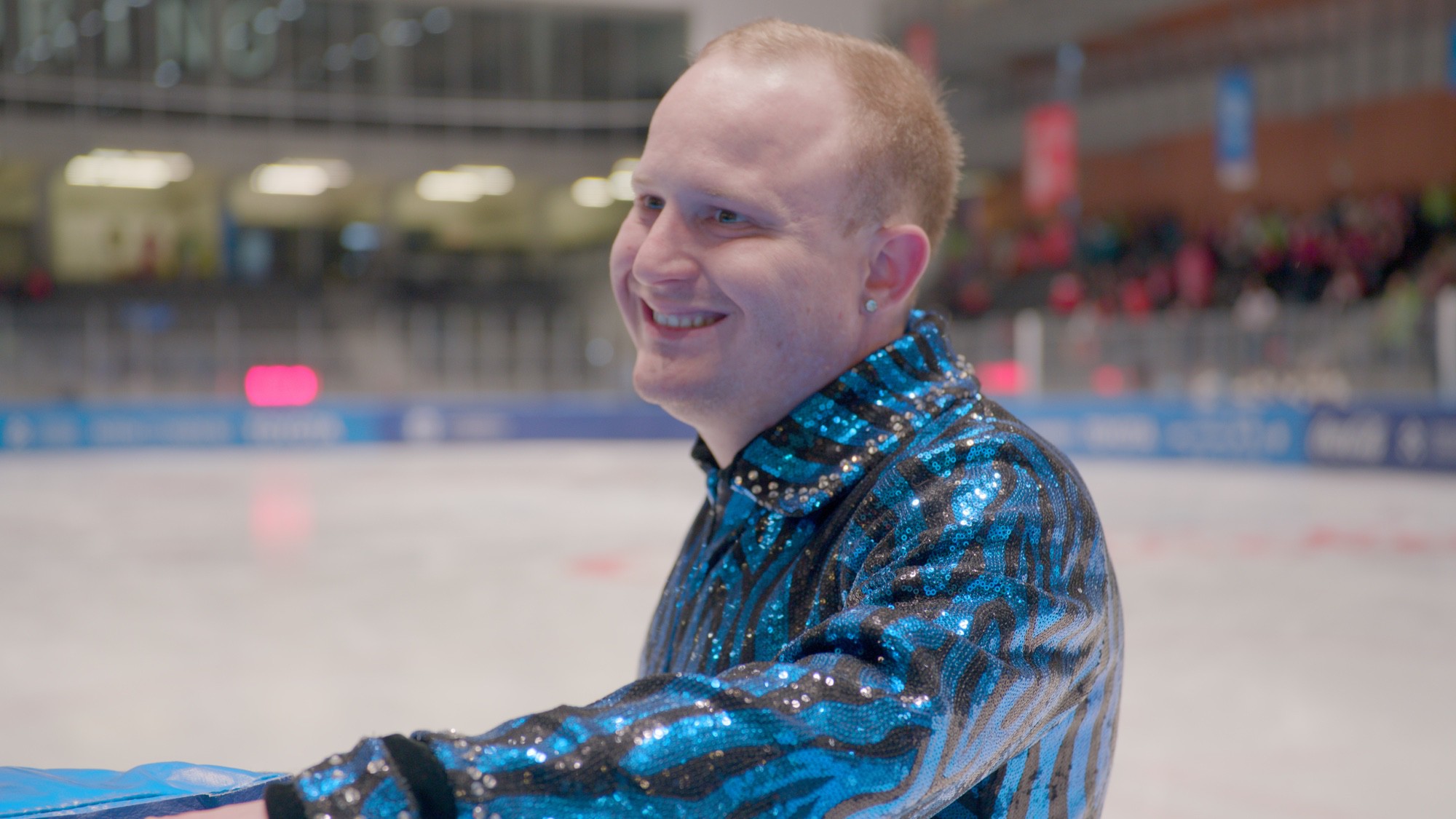 A person in a blue and black sequined jacket stands on an ice rink, smiling and holding the rink barrier.