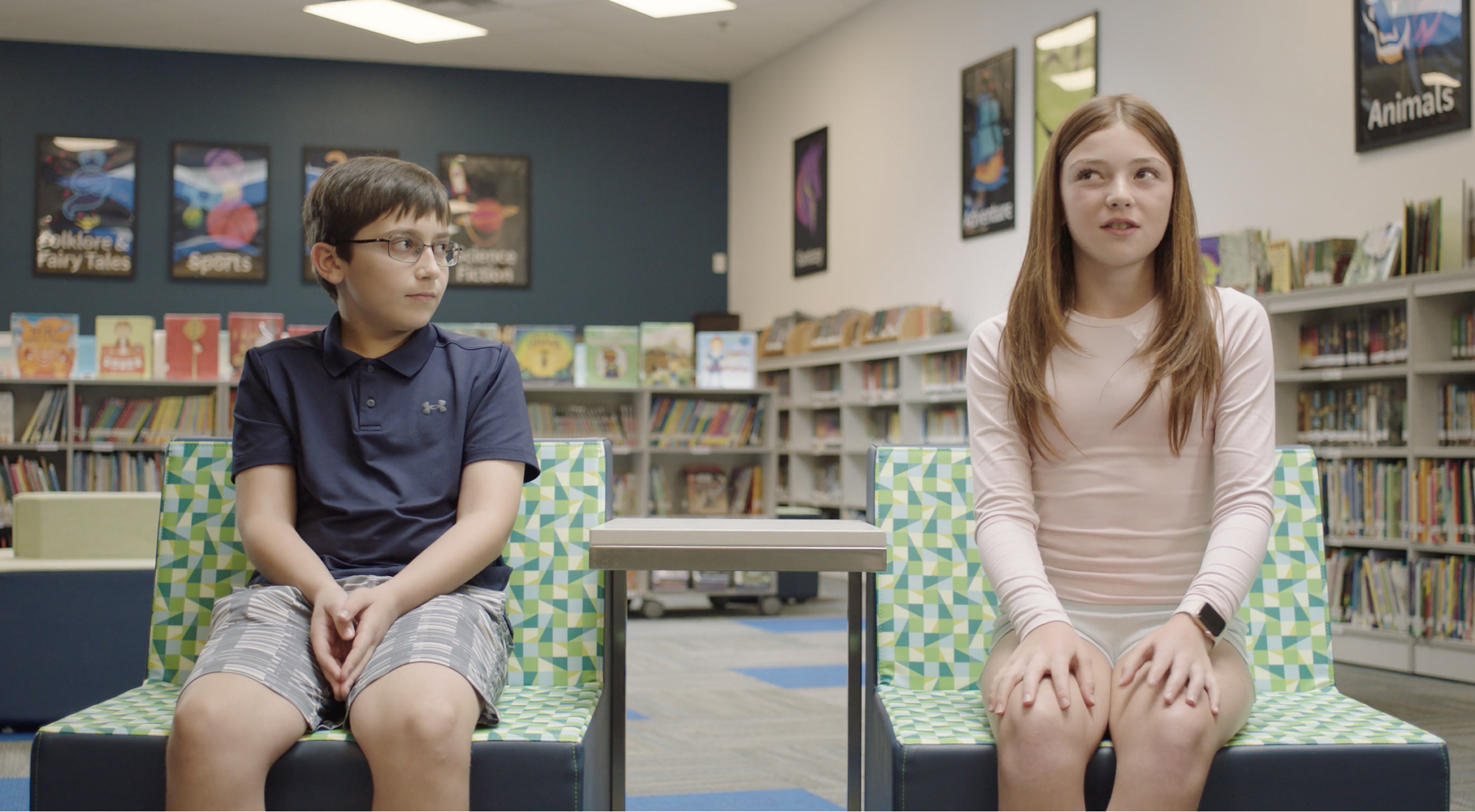 Two children sit on patterned chairs in a library, separated by a small table, with bookshelves and posters visible in the background.