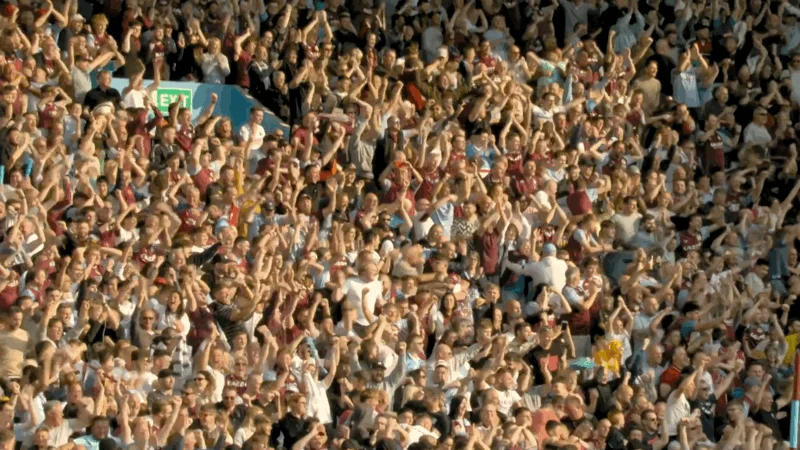 A large crowd of people in stadium seats cheer and raise their arms, suggesting a lively and energetic atmosphere at a sports event.