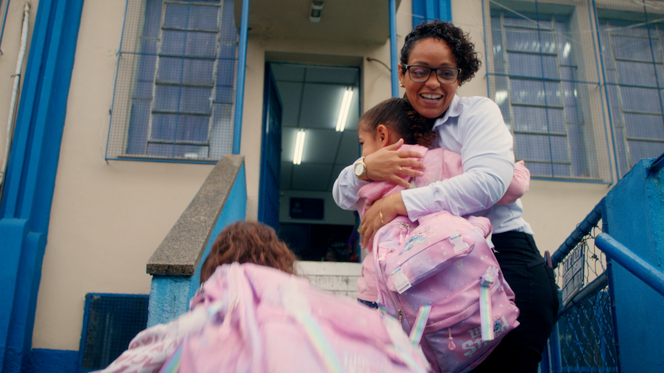 A woman smiles and hugs two children wearing pink backpacks outside the entrance of a blue and white building.