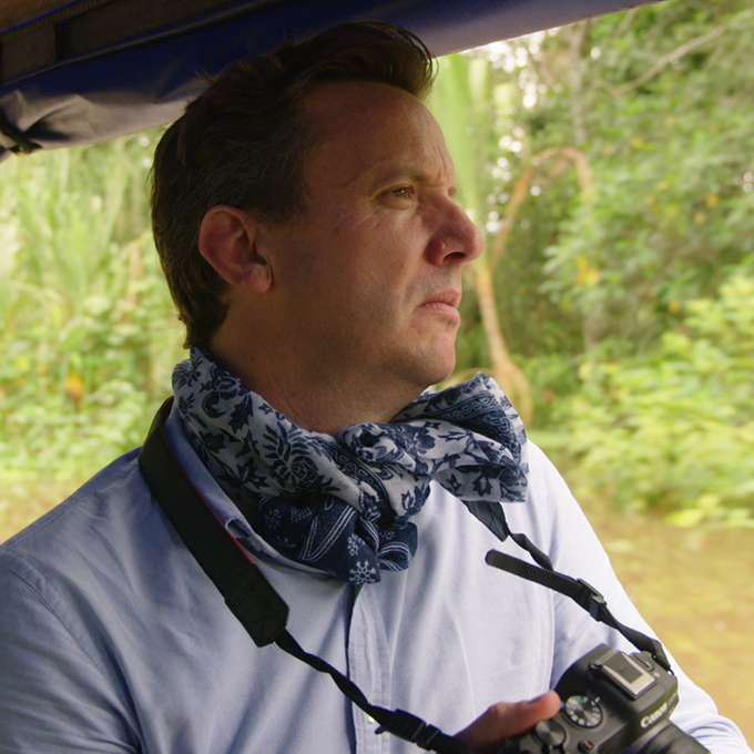 A man wearing a blue shirt and patterned scarf holds a camera while gazing out from a boat in a lush, green, riverside setting.