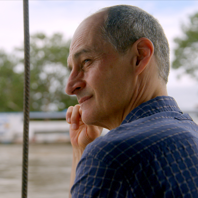 A middle-aged man with short hair and a blue checkered shirt looks thoughtfully into the distance, resting his head on his hand near an outdoor waterfront setting.