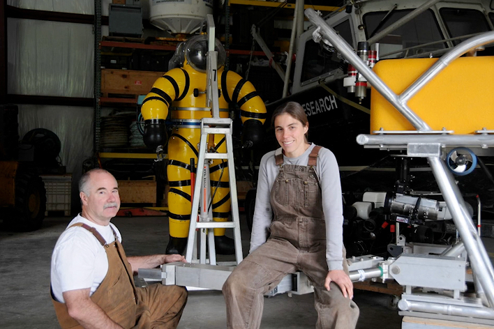 Two people in work overalls pose in a workshop next to a yellow atmospheric diving suit and underwater research equipment.