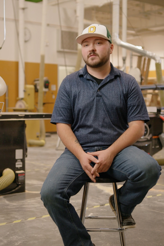A man in a blue polo shirt and jeans sits on a stool in a workshop with woodworking equipment in the background.