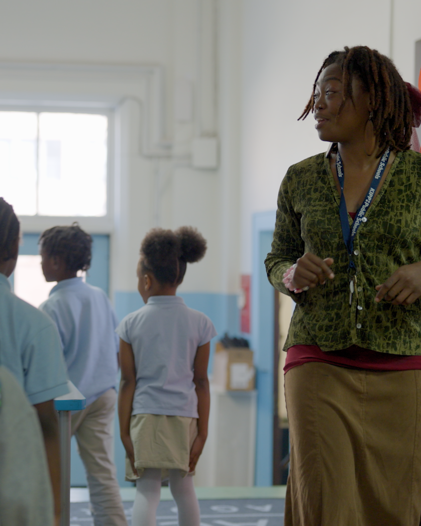 A woman with dreadlocks, wearing a green top and brown skirt, stands and gestures while several young children in school uniforms walk nearby in a brightly lit classroom.