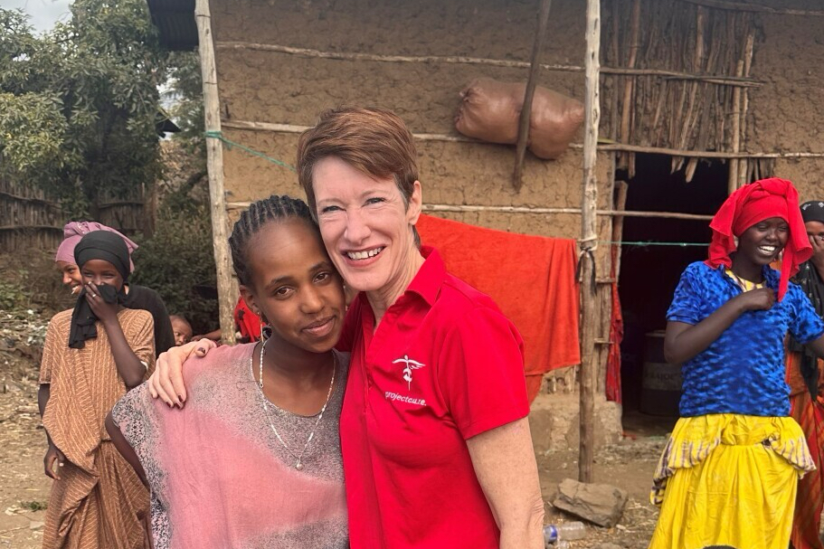 A smiling woman in a red shirt hugs a smiling young girl outdoors, with another girl standing in the background near a rustic building.