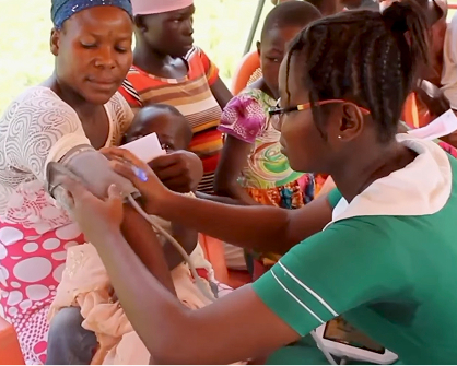 A healthcare worker in a green uniform checks a childs blood pressure while the child sits on a womans lap. Other women and children are seated in the background, likely waiting for medical attention.