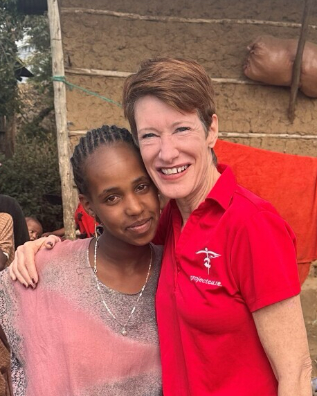 A smiling woman in a red polo shirt hugs a smiling young girl outdoors. Several people and a building appear blurred in the background.