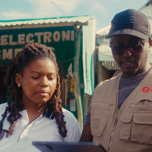 Two people stand outdoors looking at a tablet. The person on the left has long braids and wears a white shirt; the person on the right wears glasses, a cap, and a beige vest. A green electronic shop sign is visible behind them.