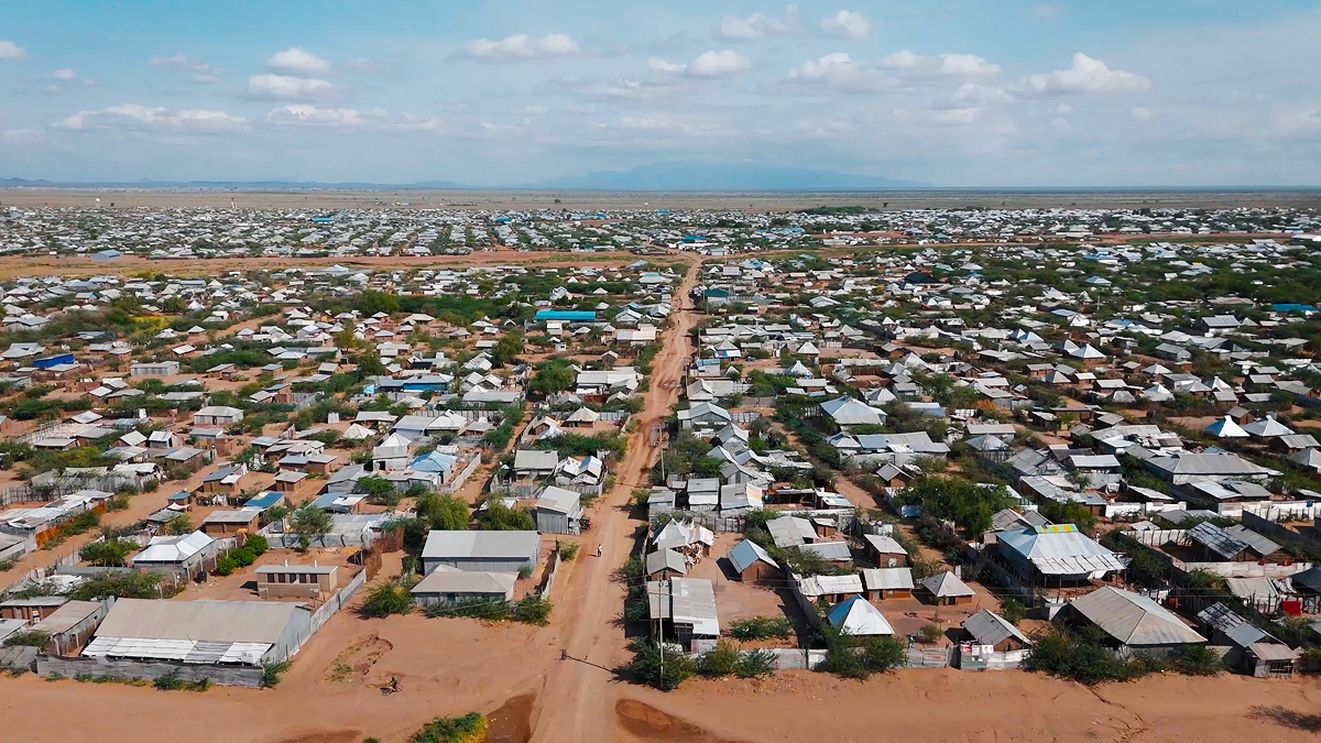 Aerial view of a sprawling refugee camp with rows of small, makeshift shelters and buildings, separated by a dirt road, under a partly cloudy sky in a dry, arid landscape.