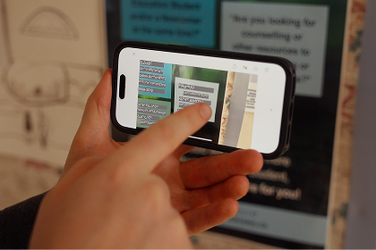 A person holds a smartphone and uses their finger to scroll through text messages displayed on the screen. A blurred sign or poster is in the background.