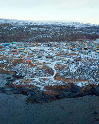 Aerial view of a small coastal town with colorful houses scattered across a rocky, snow-dusted landscape, bordered by water and rolling, barren hills in the background.