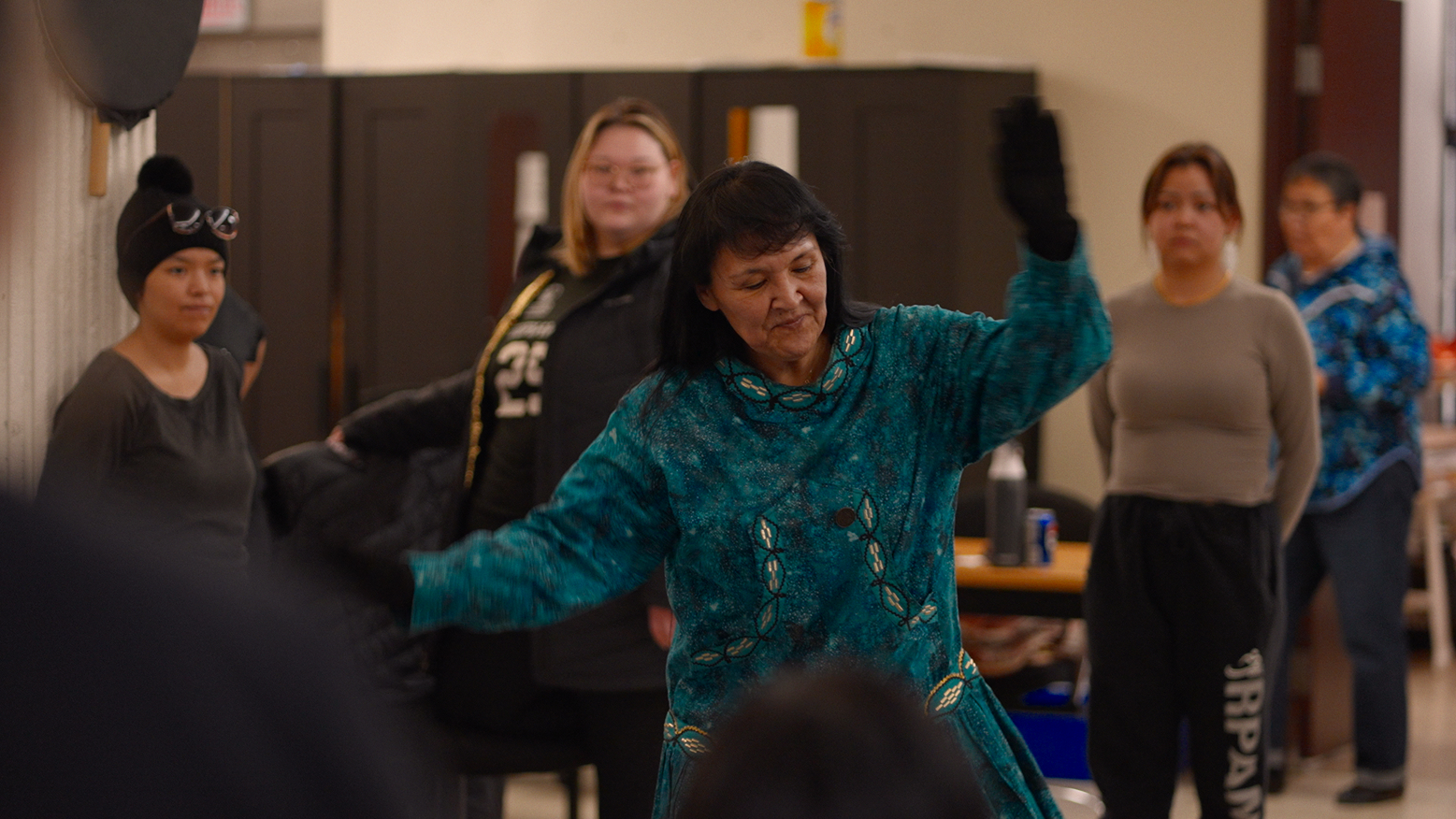 A woman in a blue patterned dress dances with one hand raised, while several people stand and watch her in a room with lockers and coats in the background.