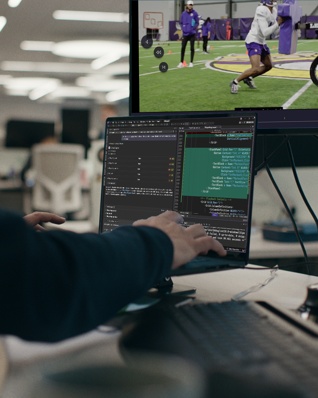 A person works at a desk with two monitors: one displaying programming code and the other showing a sports video featuring athletes training on a field.