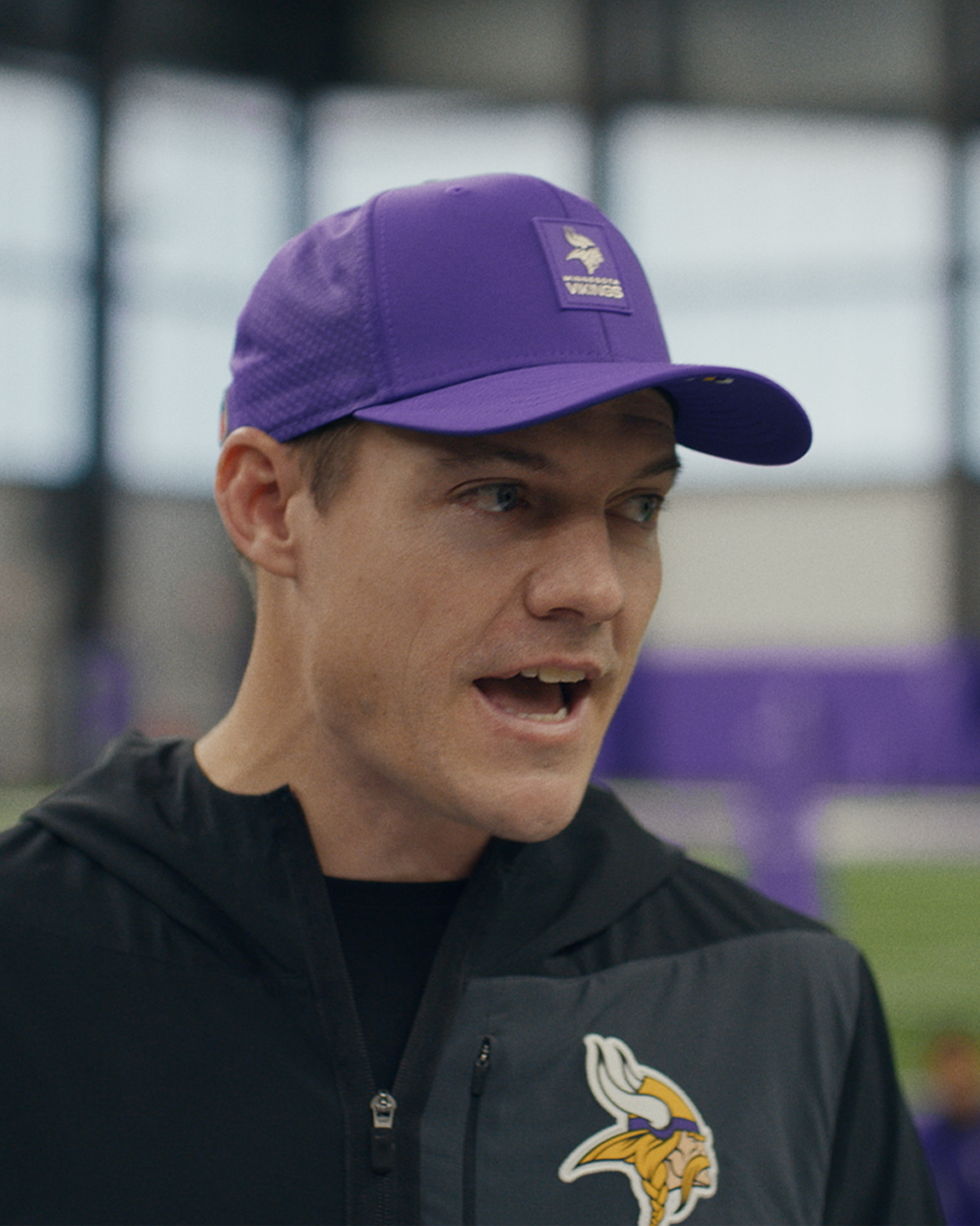 A man wearing a purple cap and a black jacket with a Minnesota Vikings logo is speaking indoors, with a blurred background of a sports facility.